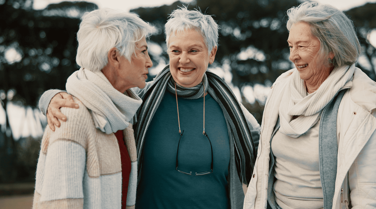 Three older women happy gynecologist accepts medicare with gray hair smiling and embracing outdoors in cool-weather clothing.
