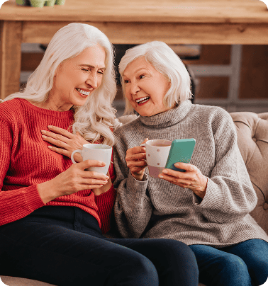 Two elderly women from Salt Lake City on medicare sitting on a couch, smiling and holding coffee mugs, with one woman holding a smartphone.