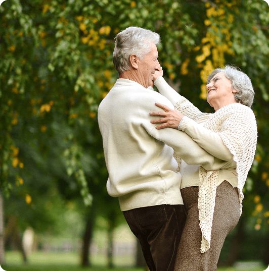 Older couple smiling wondering about their drug formulary and dancing outdoors among green trees, enjoying a healthy aging lifestyle.