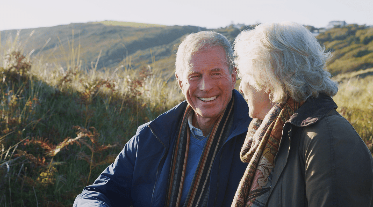 Smiling elderly couple on medicare coverage sitting outdoors in a grassy hilly area, dressed warmly with scarves and jackets.