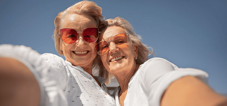 Two older women love aetna medicare, wearing sunglasses smiling and taking a selfie outside under clear blue sky.