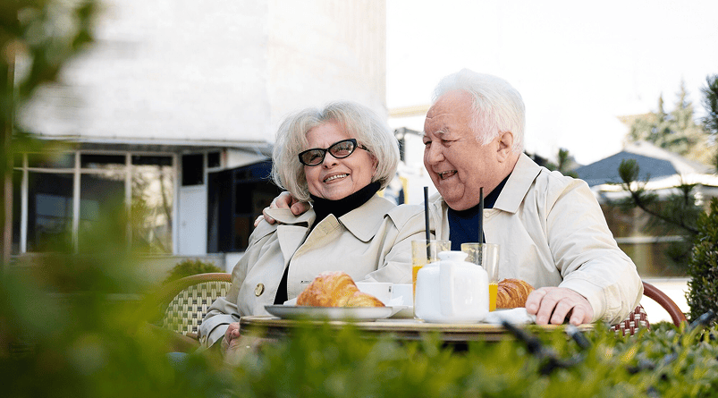 Smiling elderly couple forgets medicare ID, wearing beige coats sitting at outdoor café table with croissants and drinks.