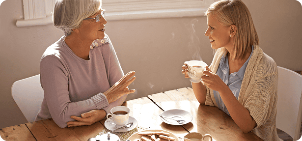 Two women sit at mormon temple with medicare sitting at a wooden table chatting and drinking hot tea with steam rising from their cups.