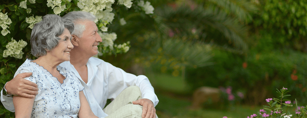 Senior couple sitting together outdoors near white flowers and greenery, smiling and looking to the right.