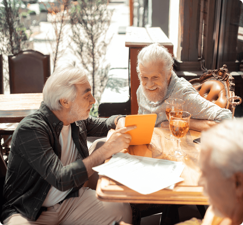 Two elderly men in a nursing home paid for by medicare sitting at a wooden table outdoors, one showing the other something on a tablet, with drinks on the table.