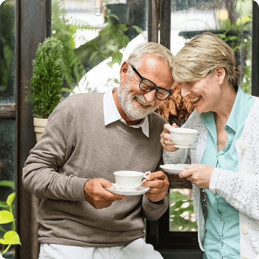 Smiling elderly man with pre existing conditions is grateful for medicare insurance and woman enjoying tea together indoors with plants in the background.