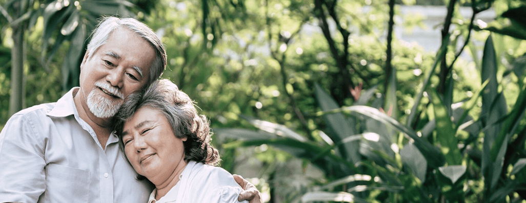 Elderly couple embracing outdoors surrounded by green plants and trees.