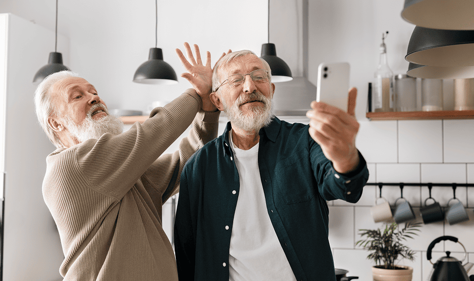 Two elderly men worried medication won't be covered by medicare insurance, in a kitchen, one taking a selfie while the other playfully raises his hand behind his head.