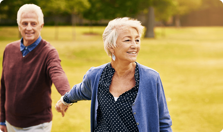 Smiling elderly couple turning 65 holding hands and walking in a sunlit park.