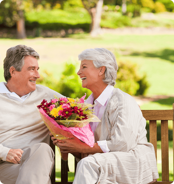 Smiling elderly couple disappointed at their doctor for not accepting medicare sitting on a bench outdoors holding a colorful bouquet of flowers.