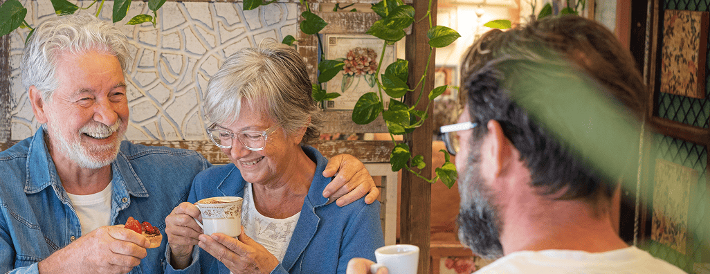 Smiling older couple enjoying coffee and pastries with a younger man in a cozy, plant-decorated setting.