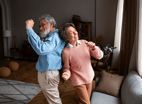 Senior couple joyfully dancing on news that their healthcare is not leaving the state of Pennsylvania, back-to-back in a cozy living room with natural light.